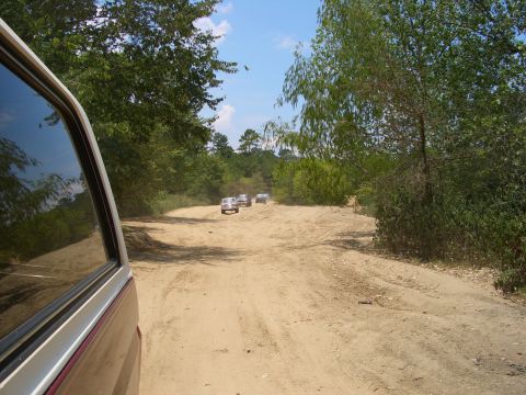 1990 Ford Bronco - A group of vehicles travels down a dusty, unpaved road surrounded by trees under a blue sky.