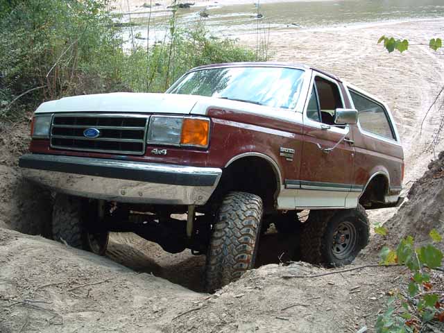 Red and white Ford Bronco with large tires stuck in a dirt ditch
