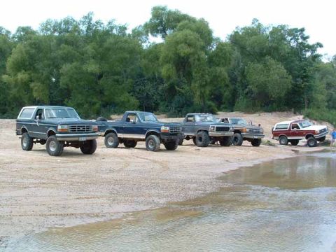 1993 Ford Bronco - Several Ford trucks and a Bronco are parked on a sandy area next to a body of water.