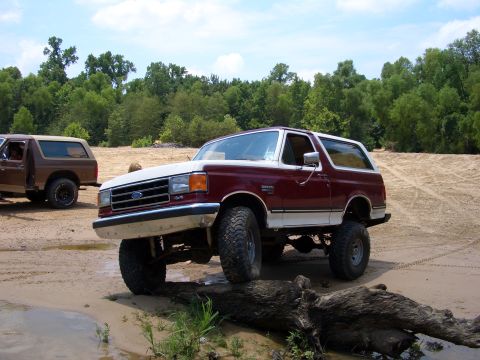1990 Ford Bronco - Two Ford Broncos are parked on a sandy area with trees in the background.