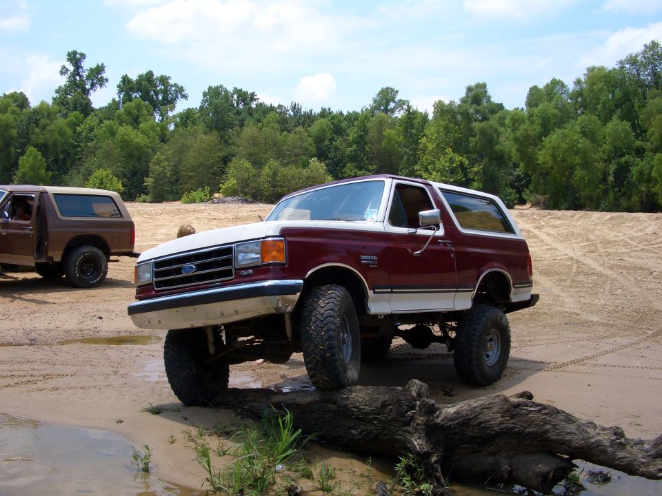 Maroon and white Ford Bronco with lifted suspension on a dirt path, front-left tire on a log