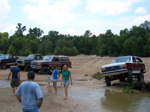 1990 Ford Bronco - A group of people are gathered around several off-road vehicles near a body of water.