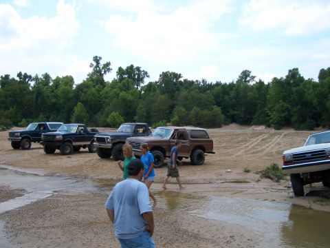 1990 Ford Bronco - A group of people walk through a muddy area near several parked trucks and SUVs, including a brown 1990 Ford Bronco.