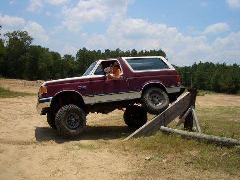 1990 Ford Bronco - A maroon and white 1990 Ford Bronco navigates an off-road obstacle on a sunny day.