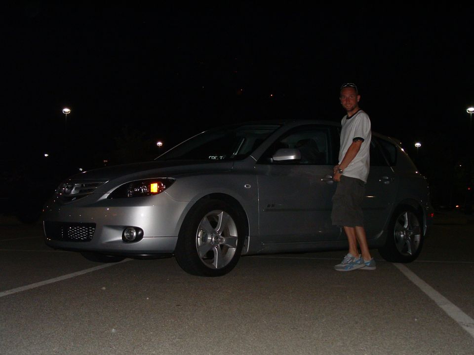 Man standing next to a silver Mazda 3 hatchback. The car has a mesh grille and distinctive headlights