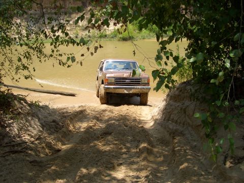 1990 Ford Bronco - A brown 1990 Ford Bronco navigates a sandy track emerging from a muddy river.