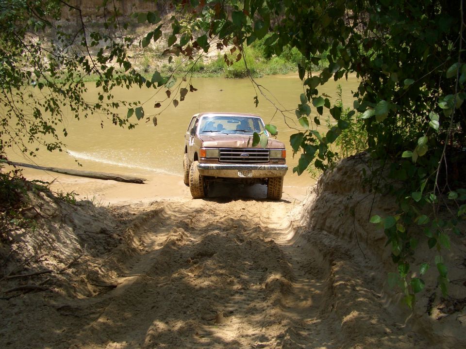 Brown 1990 Ford Bronco driving out of a muddy river on a sandy track