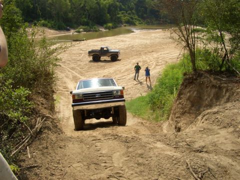 1990 Ford Bronco - A white Ford F-Series truck navigates a sandy trail near a river, with another truck visible in the background.