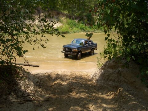 1990 Ford Bronco - Ford Bronco navigates a river crossing surrounded by lush greenery.