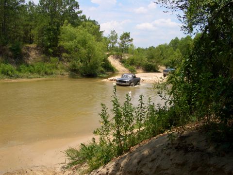 1990 Ford Bronco - Two trucks navigate a shallow riverbed surrounded by trees and sandy banks under a partly cloudy sky.