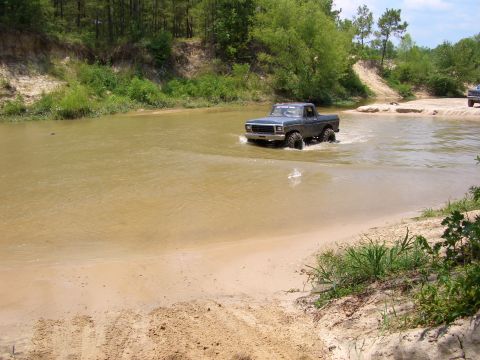 1990 Ford Bronco - A dark gray pickup truck navigates a muddy river crossing on a sunny day.