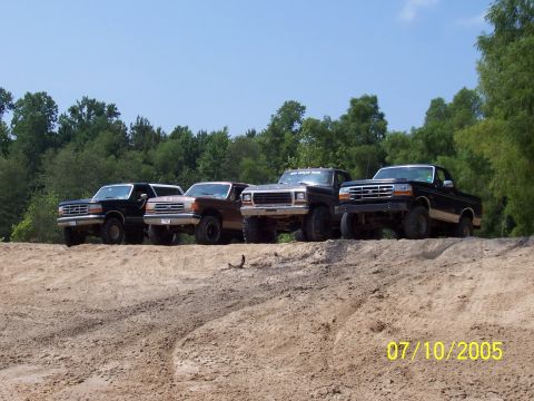 1987 Ford Bronco - Four pickup trucks are lined up on a dirt embankment under a clear blue sky.