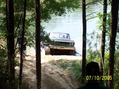 1987 Ford Bronco - Black Ford Bronco drives into a lake on a sunny day, viewed from a wooded area.