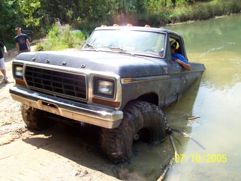 1987 Ford Bronco - A dark Ford truck with oversized tires is partially submerged in muddy water on a sunny day.
