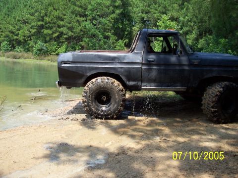 1987 Ford Bronco - A black 1978 Ford Bronco with off-road tires is partially submerged in water near a wooded area.