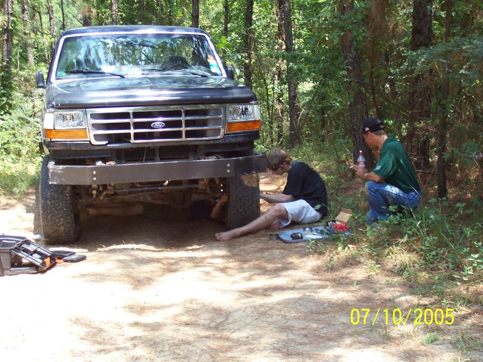 Dark gray Ford truck with aftermarket bumper, front view, with two men working on it in a wooded area