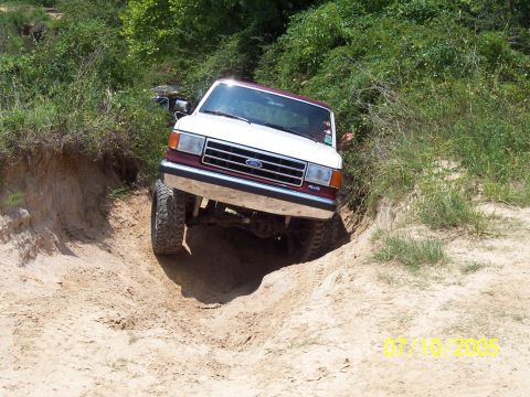 1987 Ford Bronco - This Ford Bronco is navigating a challenging off-road path with steep sandy inclines and dense vegetation.