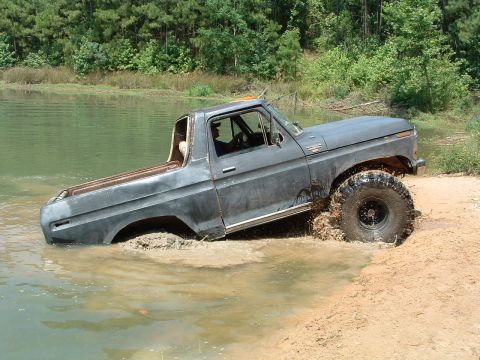 1993 Ford Bronco - A gray pickup truck navigates through water and mud, kicking up spray as it drives off a dirt embankment.