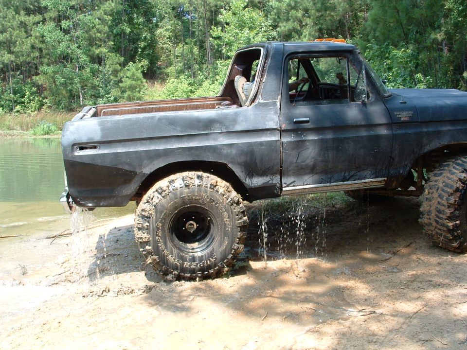 Old black Ford Bronco with large tires stuck in mud near water