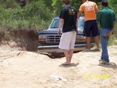 1987 Ford Bronco - A dark-colored Ford Bronco is partially submerged in a dirt ditch, with three men standing nearby.