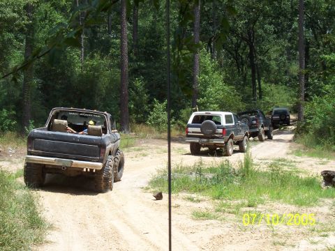 1987 Ford Bronco - A convoy of off-road vehicles navigates a dusty trail surrounded by dense forest foliage.