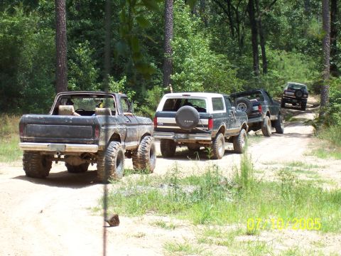 1987 Ford Bronco - Multiple Ford Broncos drive down a dirt road in a wooded area.