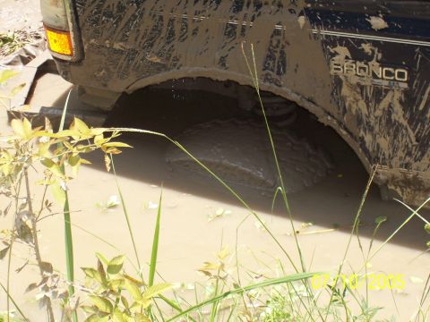 1987 Ford Bronco - A Ford Bronco is stuck in muddy water, with the rear wheel deep in the mire.