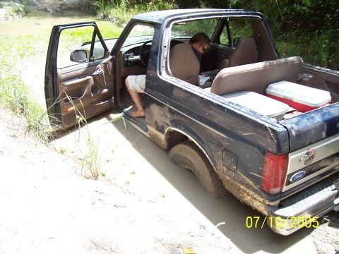 1987 Ford Bronco - A 1987 Ford Bronco is stuck in the mud with the driver's door open and a person exiting.