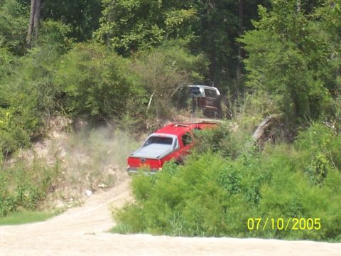 1987 Ford Bronco - A red pickup truck kicks up dust as it drives on a dirt road, with another vehicle partially visible behind it.