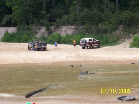 1987 Ford Bronco - Two off-road vehicles are parked on a sandy bank next to a body of water.