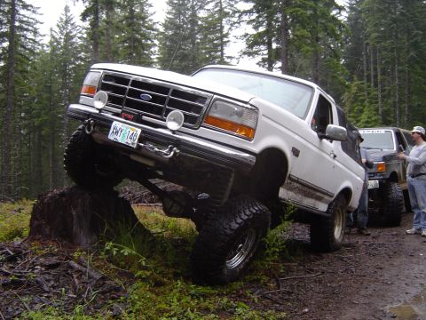 1989 Ford Bronco - A white 1989 Ford Bronco navigates an obstacle during an off-road event, with another vehicle and participants visible.