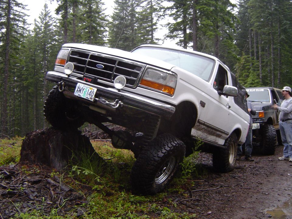 White lifted Ford Bronco climbing a tree stump with its front tire on a forest trail