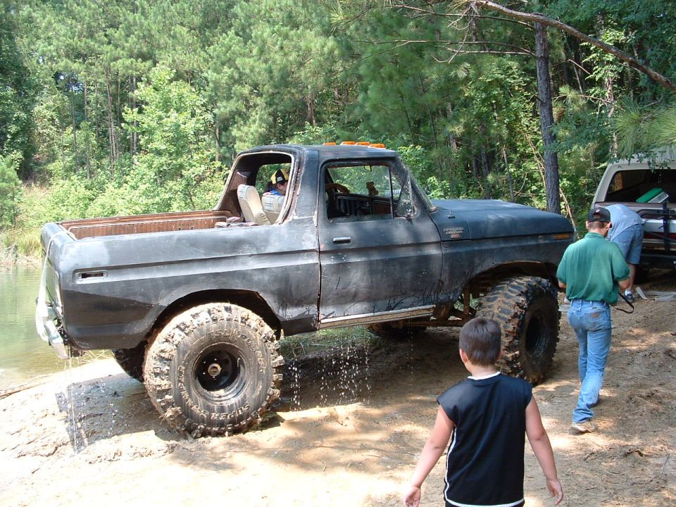 Modified Ford Bronco with large tires on a dirt path near water, another vehicle behind it