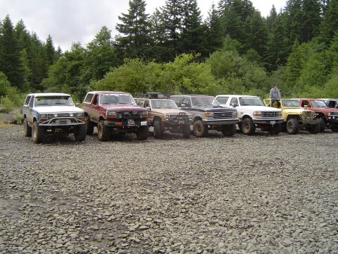 1989 Ford Bronco - Several off-road vehicles are lined up on a gravel lot with trees in the background.