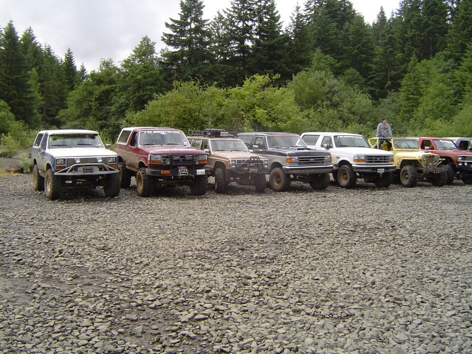 Row of lifted Ford Broncos, a Nissan pickup and Jeeps parked on a rocky staging area by a forest