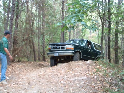 1993 Ford Bronco - A 1993 Ford Bronco navigates an off-road trail surrounded by trees, with a person watching from the side.
