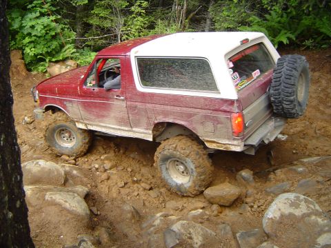 1989 Ford Bronco - A red and white Ford Bronco tackles a challenging off-road course with mud and rocks.