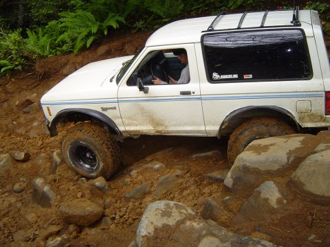 1989 Ford Bronco - A white 1989 Ford Bronco tackles a challenging off-road course with large rocks and mud.