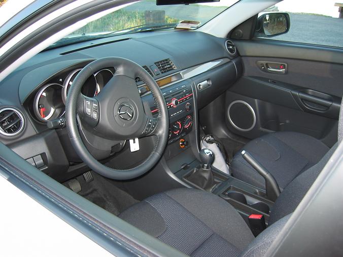 Car interior showing dashboard, steering wheel with Mazda logo, center console with gear shift, and seats