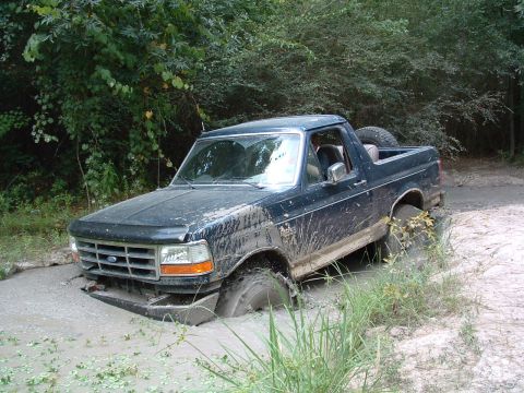1993 Ford Bronco - A dark blue Ford Bronco is stuck deep in mud, with only its rear half visible above the mire.