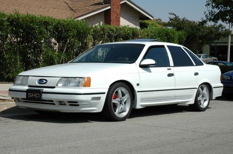 1993 Ford Taurus - A white 1993 Ford Taurus SHO is parked on a residential street with a house and bushes in the background.