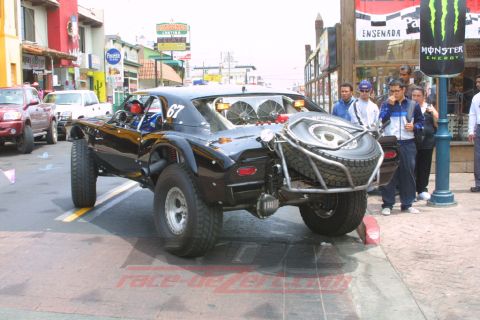 1990 Ford Bronco - black off-road race car marked number 67 with a rear-mounted spare tire sits on a city street as onlookers watch.