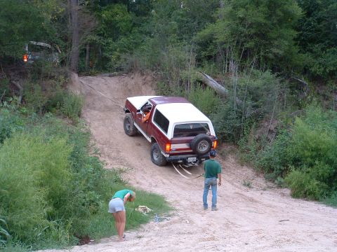 1993 Ford Bronco - A red and white Ford Bronco is being towed up a steep dirt incline by a tow strap, with assistance from another vehicle out of frame.