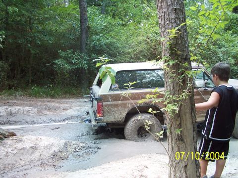 1987 Ford Bronco - A brown 1987 Ford Bronco is stuck in a muddy trail, with a young boy observing the scene.