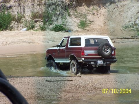 1987 Ford Bronco - A maroon and white Ford Bronco navigates shallow water with a spare tire on its rear.