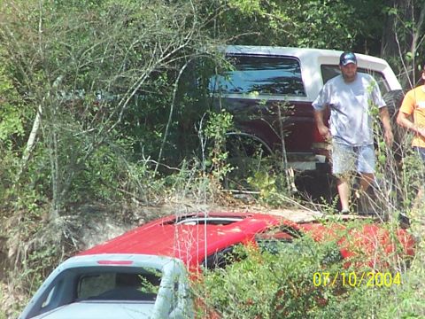 1987 Ford Bronco - A maroon and white Ford Bronco is seen off-road, with a red car in front of it and people standing nearby.