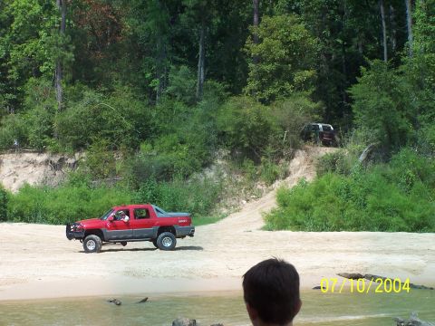 1987 Ford Bronco - Two vehicles are visible in a wooded, sandy area near water.
