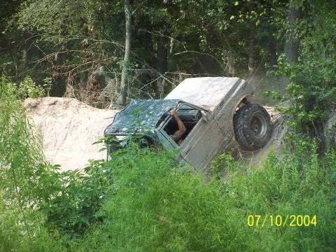 1987 Ford Bronco - Off-road vehicle stuck on a muddy embankment, surrounded by dense vegetation.