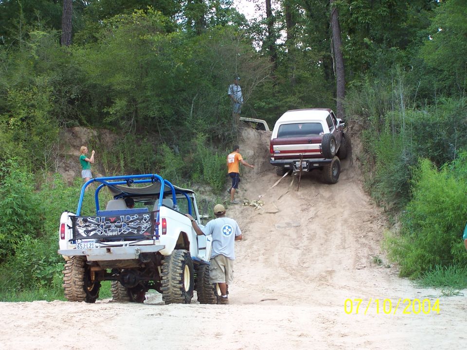 Two off-road vehicles, one white with a blue roll cage and one maroon and white SUV, ascend a dirt hill with people around