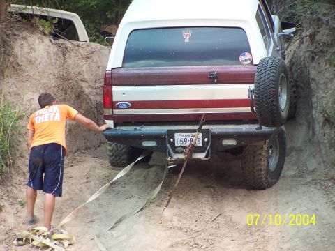 1987 Ford Bronco - A 1987 Ford Bronco is seen from the rear, stuck in a dirt path with a person assisting. Another vehicle is visible in the background.